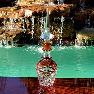 May include: A clear and amber-colored glass decanter featuring a tall, pointed stopper. The body of the decanter displays intricate cut-glass patterns, including a band of etched floral motifs and geometric diamond shapes around the base.