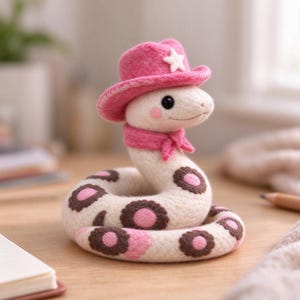 Könnte beinhalten: Eine nadelgefilzte, cremefarbene Schlangenfigur mit einem rosa Cowboyhut mit weißem Stern und einem passenden rosa Schal. Die Schlange hat dunkelbraune und rosa kreisförmige Flecken auf ihrem Körper, schwarze Knopfaugen und rosa Wangenakzente.