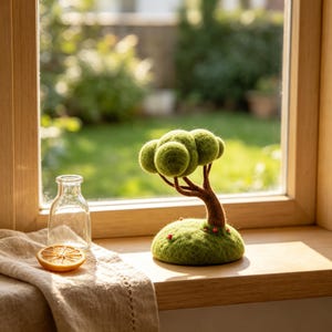 May include: A needle-felted green tree sculpture with a brown trunk, resting on a rounded green base featuring tiny red accents. Beside the tree sits a clear glass bottle and a single dried orange slice on a textured beige fabric cloth.