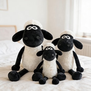 May include: Three plush sheep toys featuring black faces, ears, and limbs with white woolly bodies. Each sheep has large, round white eyes with black pupils. The toys are arranged in a group, with one large sheep on each side and a smaller one in the centre.