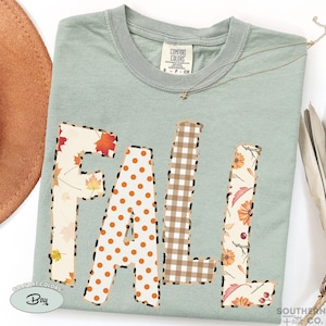 May include: A sage green t-shirt featuring the word FALL in large block letters. Each letter has a unique pattern: F has autumn leaves, A has orange polka dots, L has brown and white gingham, and L has floral motifs. All letters have a leopard print border.