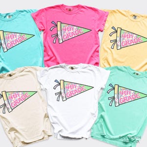 May include: A collection of six short-sleeve crew neck t-shirts in various colors including pink, white, yellow, teal, and light green. Each shirt features a graphic of a pennant flag with the text 4th GRADE in pink letters.