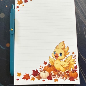 May include: A white lined notepad featuring a yellow cartoon bird character, orange and white pumpkins, and scattered autumn maple leaves in red, orange, and yellow colors. A solid teal retractable ballpoint pen is placed beside the notepad.