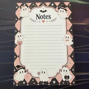 May include: A rectangular notepad featuring a black and light pink diamond pattern border with white stars. The center has a white lined area with the word Notes at the top, decorated with a bat, a heart, and six cartoon ghosts holding Halloween items.
