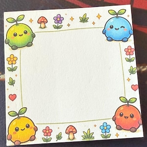 Peut inclure: Un bloc-notes carré blanc avec une bordure décorative illustrée de petites créatures rondes, de champignons, de fleurs, de feuilles, de cœurs et d'étoiles scintillantes. Le centre de la note est vide pour écrire des messages ou des notes.
