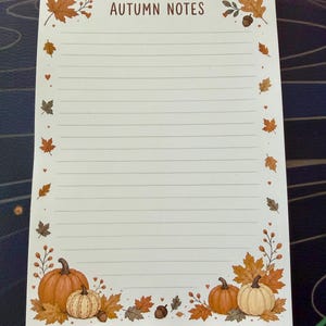 May include: A rectangular notepad with white paper featuring horizontal lines and the text AUTUMN NOTES at the top. The border is decorated with illustrations of orange and cream pumpkins, brown acorns, and various autumn leaves in shades of brown.
