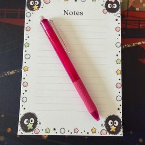 May include: A rectangular notepad with white lined paper featuring the word Notes at the top, decorated with small black fuzzy creatures and colorful stars along the border. A bright pink retractable ballpoint pen rests diagonally on the paper.