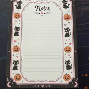 May include: A rectangular notepad with a white lined center and the word Notes printed at the top. The border features a black background with repeating illustrations of black cats, orange jack-o'-lanterns, pink hearts, and stars, framed by a light pink scalloped edge.