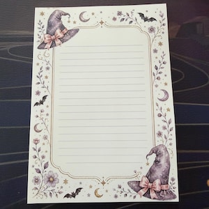 May include: A rectangular sheet of lined stationery featuring a decorative border with purple witch hats, pink bows, crescent moons, stars, small flowers, and black bats. The central area is white with horizontal lines for writing text.