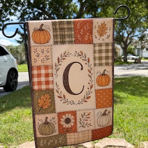 May include: A rectangular garden flag featuring a patchwork design of autumn-themed squares. The center panel displays a large letter C surrounded by a wreath of leaves. The surrounding squares include illustrations of pumpkins, sunflowers, and foliage.