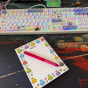 May include: A rectangular notepad with lined paper featuring a border of colorful cartoon creatures, flowers, and mushrooms, with the word Notes printed at the top. A bright pink retractable ballpoint pen rests diagonally across the notepad.