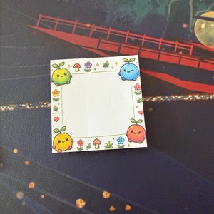 May include: A square sticky note featuring a white center bordered by a thin gold line. The border is decorated with small colorful flowers, mushrooms, and four round, smiling fruit-like characters in green, blue, orange, and yellow.