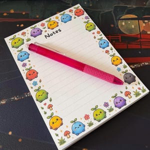 May include: A white rectangular notepad with horizontal lines and the word Notes printed at the top. The border features colorful cartoon characters with sprouts, mushrooms, and flowers. A bright pink retractable ballpoint pen rests on top.