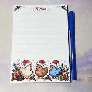 Peut inclure: Un bloc-notes ligné blanc avec le mot Notes imprimé en haut, décoré de houx et de flocons de neige. Le bas présente trois poulets en pixel art portant des bonnets de Père Noël, entourés de cadeaux, de sucres d'orge et de houx. Un stylo bleu.