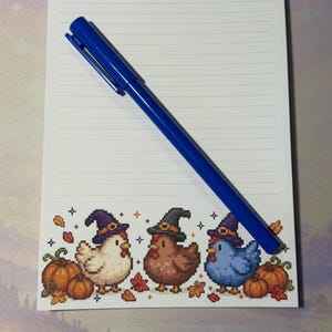 Peut inclure: Un bloc-notes ligné blanc avec le mot Notes en haut et une illustration en pixel art en bas représentant trois poulets portant des chapeaux de sorcière entourés de citrouilles et de feuilles d'automne, avec un stylo bleu posé sur le papier.