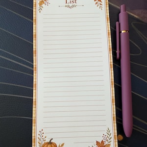 May include: A rectangular notepad with a tan plaid border, featuring the word List at the top, decorated with illustrations of orange pumpkins and autumn leaves in the corners, accompanied by a matte mauve retractable ballpoint pen.