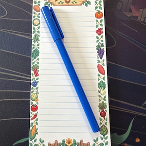 May include: A rectangular notepad with blue horizontal lines and a border featuring colorful illustrations of various vegetables, fruits, and flowers. The top header reads Shop at Pierre's. A blue plastic ballpoint pen rests on the notepad.