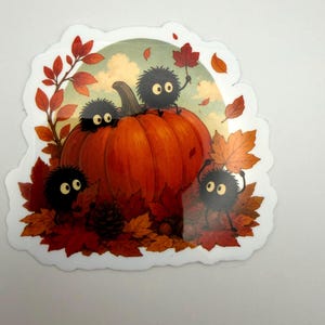 May include: A die-cut sticker featuring a large orange pumpkin surrounded by autumn leaves and a pinecone. Four small, round, black fuzzy creatures with large eyes are interacting with the pumpkin and leaves in a fall-themed outdoor setting.