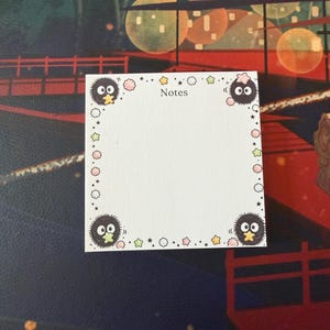 May include: A square white notepad with the word Notes printed at the top center. The border features a repeating pattern of small colorful stars, circles, and four black fuzzy soot sprite characters, each holding a small yellow or green star.