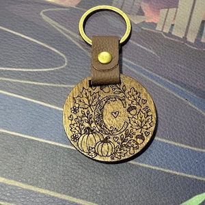 May include: A circular wooden keychain featuring a laser-engraved letter C surrounded by autumn motifs including pumpkins, maple leaves, acorns, and small hearts. The wood disc is attached to a brown leather strap with a brass rivet and a gold metal ring.