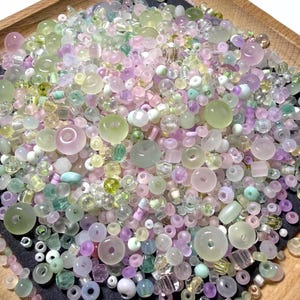 May include: A large collection of assorted loose beads in various shades of pastel pink, light green, lavender, and clear glass. The beads vary in size, shape, and texture, including round, faceted, and donut-shaped styles scattered across a dark surface.