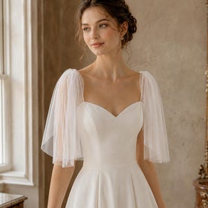 May include: A white floor-length dress featuring a sweetheart neckline and sheer, layered tulle flutter sleeves. The bodice is fitted with a clean, smooth finish, transitioning into a full, pleated skirt that flows downward from the waistline.