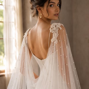 May include: A white floor-length gown featuring a deep back neckline and thin straps. Attached to the shoulders are sheer tulle cape panels adorned with clusters of white pearls at the top and scattered pearl embellishments throughout the fabric.
