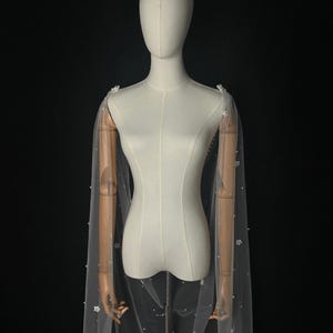 May include: A sheer white bridal cape made of tulle, featuring delicate scattered white floral appliqu&eacute;s and small white pearls throughout the fabric. The cape drapes from the shoulders, creating a long, flowing silhouette against a dark background.