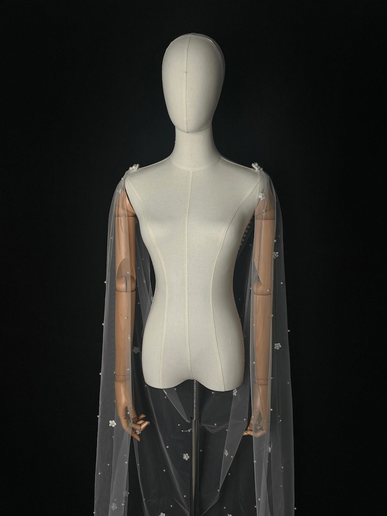 May include: A sheer white bridal cape made of tulle, featuring delicate scattered white floral appliqu&eacute;s and small white pearls throughout the fabric. The cape drapes from the shoulders, creating a long, flowing silhouette against a dark background.