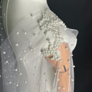 May include: A sheer white tulle capelet adorned with scattered white pearls. The shoulder features an intricate, dense floral-shaped appliqu&eacute; composed of numerous white pearls stitched onto a white fabric base, creating a textured, layered effect.