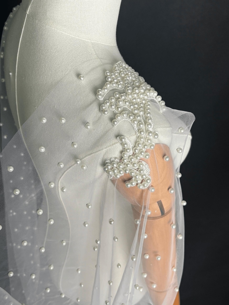 May include: A sheer white tulle capelet adorned with scattered white pearls. The shoulder features an intricate, dense floral-shaped appliqu&eacute; composed of numerous white pearls stitched onto a white fabric base, creating a textured, layered effect.
