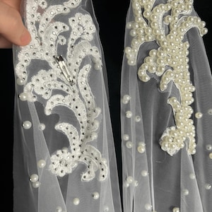 May include: Two white tulle hair accessories featuring intricate floral embroidery. One piece is embellished with small silver rhinestones and a metal hair clip, while the other is adorned with numerous white faux pearls scattered across the fabric.
