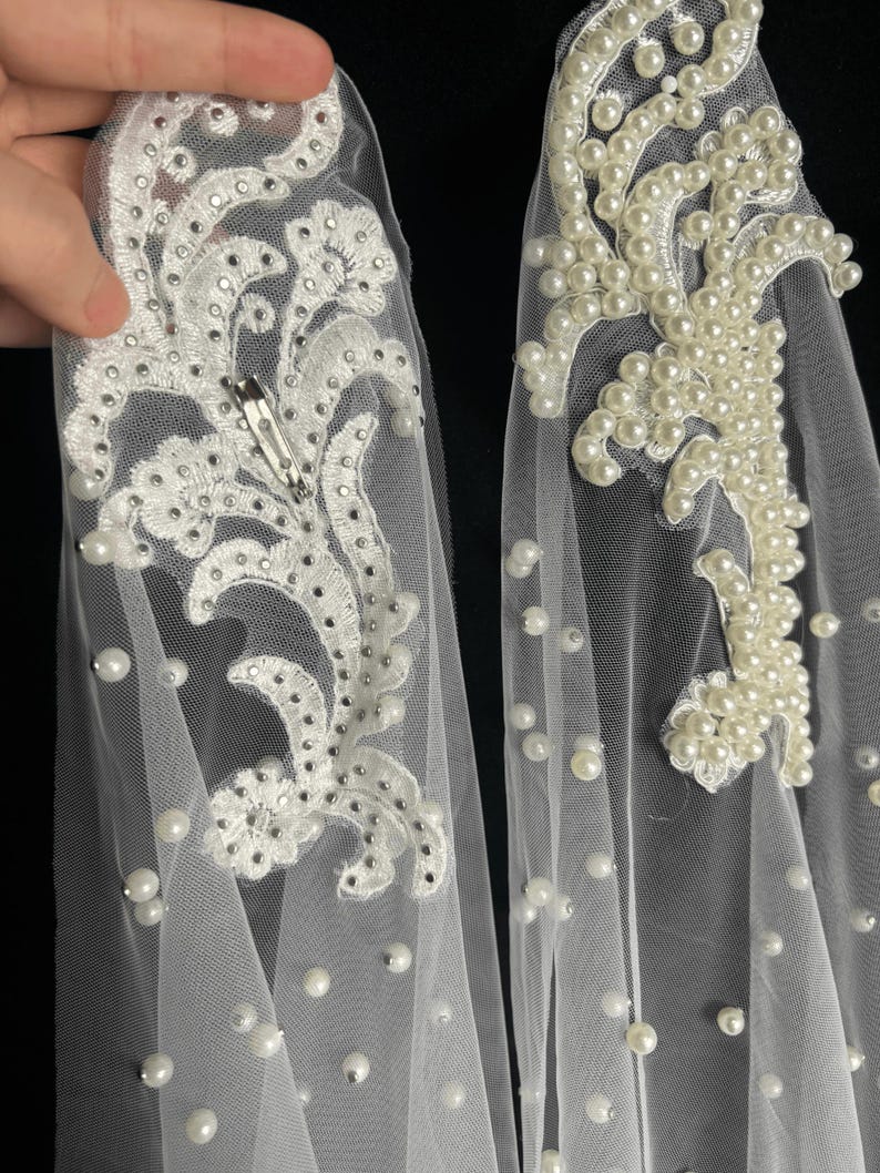 May include: Two white tulle hair accessories featuring intricate floral embroidery. One piece is embellished with small silver rhinestones and a metal hair clip, while the other is adorned with numerous white faux pearls scattered across the fabric.