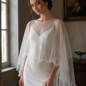 May include: A white sleeveless slip dress with a V-neckline, paired with a sheer, lightweight tulle capelet. The capelet features a delicate, wavy lettuce-edge hemline that drapes over the shoulders and arms, creating a soft, layered silhouette.