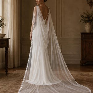 May include: A floor-length white gown featuring a deep back neckline and a long, sheer tulle cape attached at the shoulders. The cape is adorned with delicate white leaf-patterned embroidery along the edges and drapes to the floor.