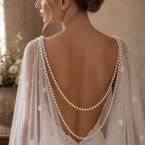 May include: A sheer white bridal cape featuring delicate floral appliqu&eacute;s and scattered pearls. The cape is adorned with two draped strands of white pearls that hang across the open back, connecting at the shoulders to create a layered effect.