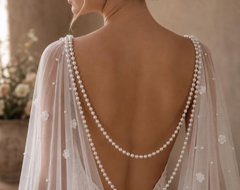 May include: A sheer white bridal cape featuring delicate floral appliqués and scattered pearls. The cape is adorned with two draped strands of white pearls that hang across the open back, connecting at the shoulders to create a layered effect.