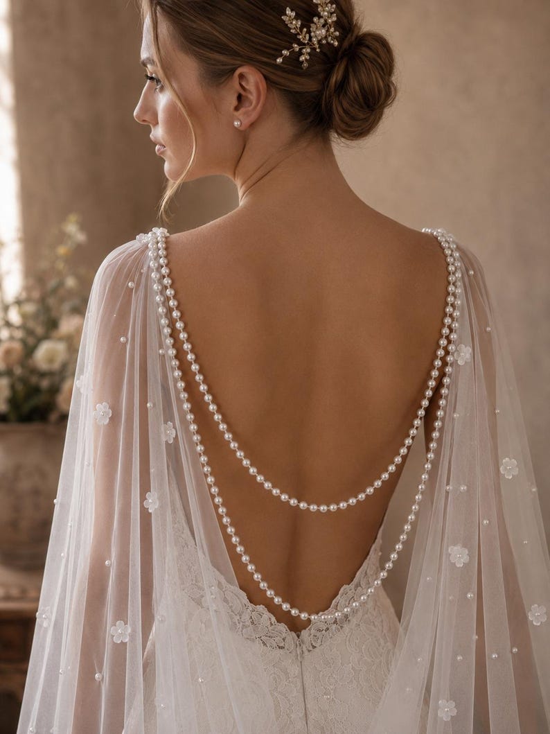 May include: A sheer white bridal cape featuring delicate floral appliqu&eacute;s and scattered pearls. The cape is adorned with two draped strands of white pearls that hang across the open back, connecting at the shoulders to create a layered effect.