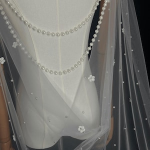 May include: A sheer white tulle veil featuring two draped strands of white pearls across the back. The fabric is adorned with scattered individual white pearls and small white floral appliqu&eacute;s, creating a delicate and layered appearance.