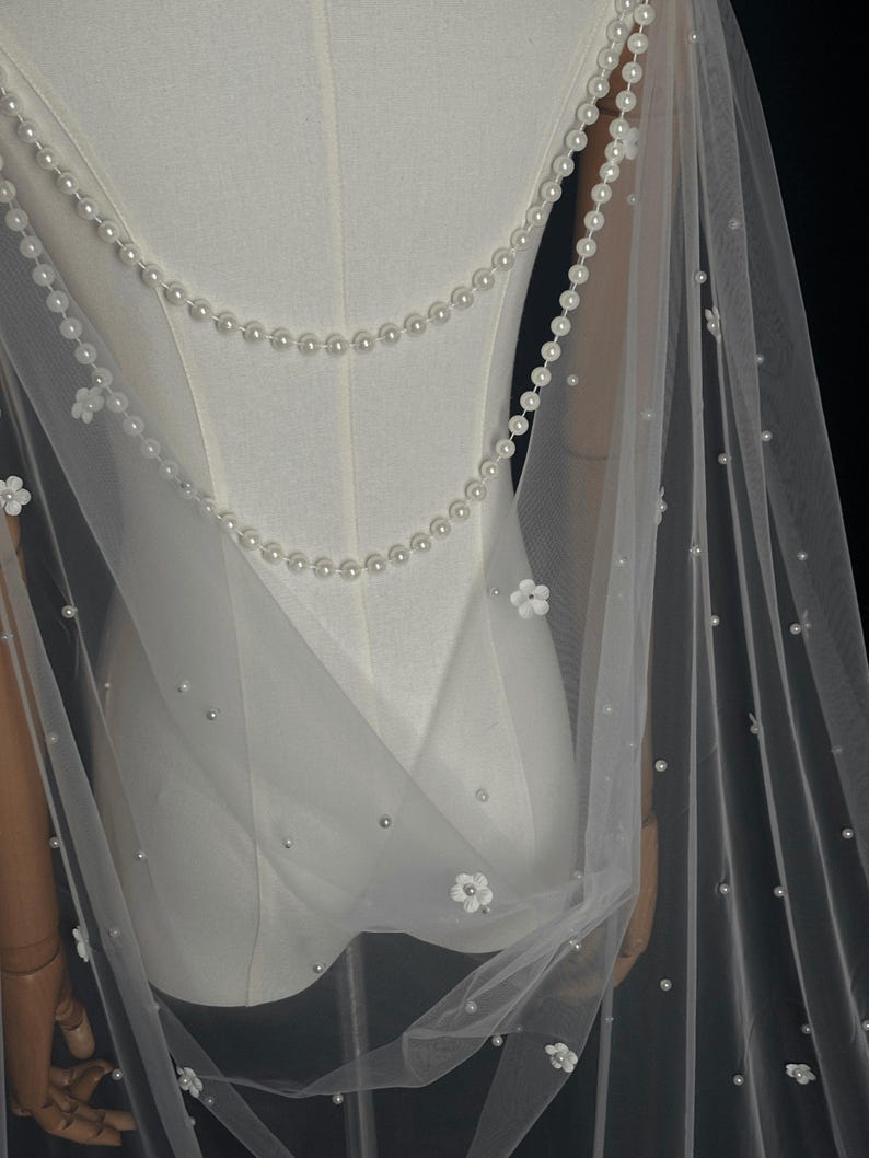 May include: A sheer white tulle veil featuring two draped strands of white pearls across the back. The fabric is adorned with scattered individual white pearls and small white floral appliqu&eacute;s, creating a delicate and layered appearance.