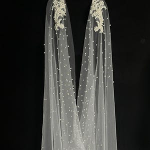 May include: A pair of long, sheer white tulle veils adorned with scattered white pearls throughout the fabric. Each veil features an intricate, elongated floral-shaped applique at the top, densely embellished with white beads and pearls.