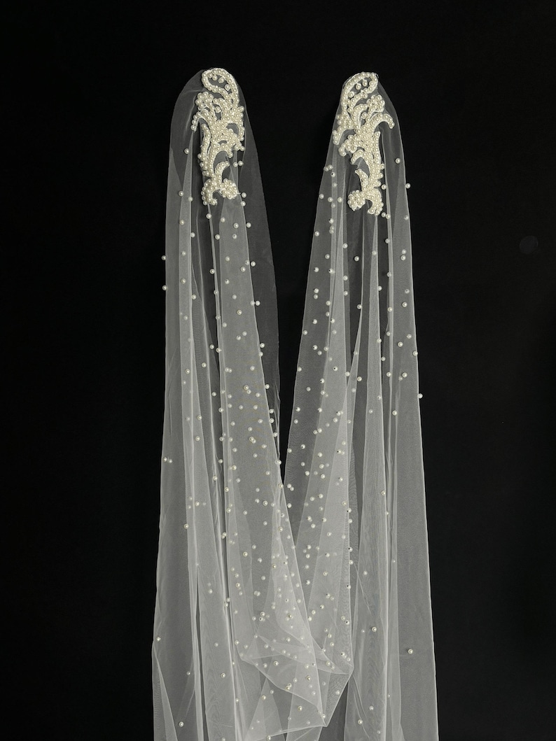 May include: A pair of long, sheer white tulle veils adorned with scattered white pearls throughout the fabric. Each veil features an intricate, elongated floral-shaped applique at the top, densely embellished with white beads and pearls.