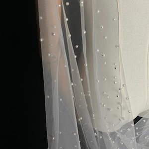 May include: A sheer white tulle veil featuring scattered round white pearls throughout the fabric. The lightweight material drapes over a mannequin, showcasing the evenly distributed pearl embellishments across the translucent, fine-mesh surface.