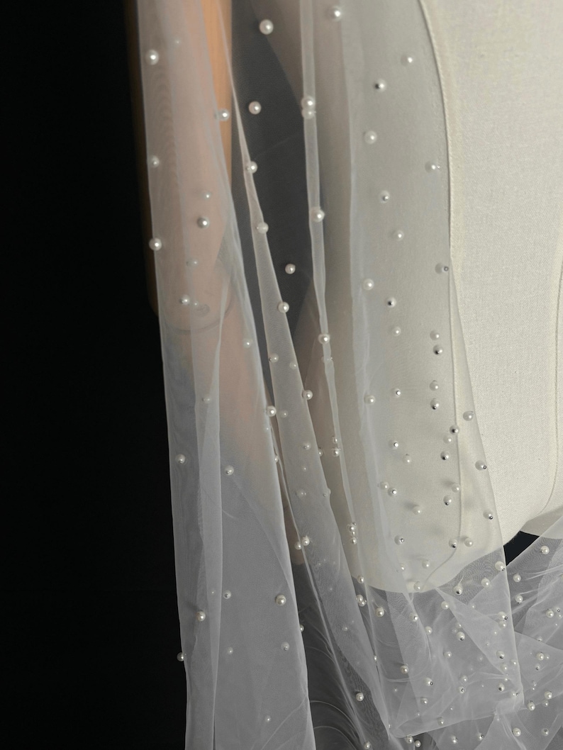 May include: A sheer white tulle veil featuring scattered round white pearls throughout the fabric. The lightweight material drapes over a mannequin, showcasing the evenly distributed pearl embellishments across the translucent, fine-mesh surface.