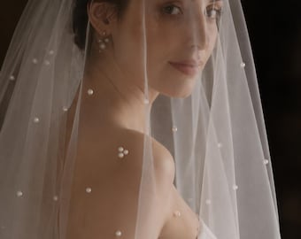 May include: A sheer white tulle wedding veil adorned with scattered small white pearls throughout the fabric. The veil is draped over the head and shoulders, featuring a delicate texture and a soft, translucent appearance that covers the upper body.