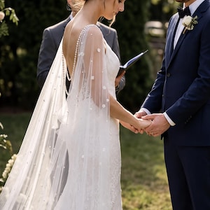 May include: A white floor-length wedding dress with a low back and a sheer cape featuring delicate pearl and floral embellishments. The dress has a lace bodice and a flowing skirt, paired with a dark navy blue suit jacket and a black bow tie.
