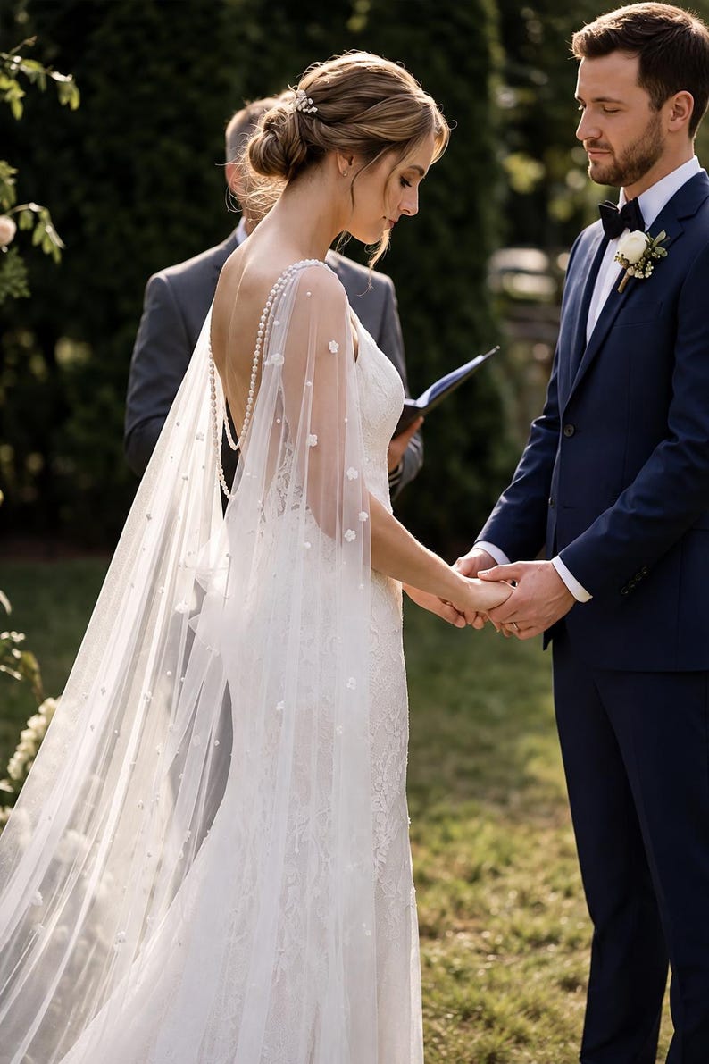 May include: A white floor-length wedding dress with a low back and a sheer cape featuring delicate pearl and floral embellishments. The dress has a lace bodice and a flowing skirt, paired with a dark navy blue suit jacket and a black bow tie.