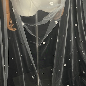 May include: A sheer white tulle veil featuring scattered small white faux pearls and delicate three-dimensional white floral appliqu&eacute;s. The lightweight fabric drapes elegantly, showcasing a consistent pattern of embellishments across the surface.