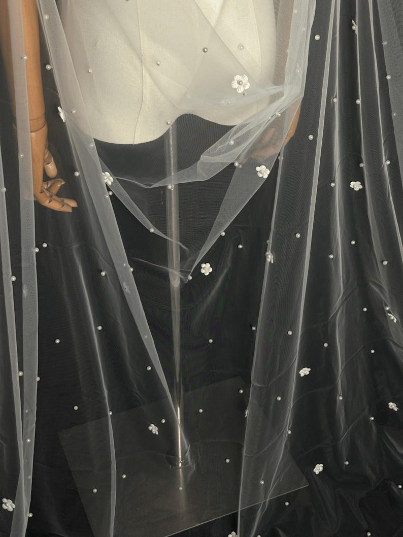 May include: A sheer white tulle veil featuring scattered small white faux pearls and delicate three-dimensional white floral appliqu&eacute;s. The lightweight fabric drapes elegantly, showcasing a consistent pattern of embellishments across the surface.