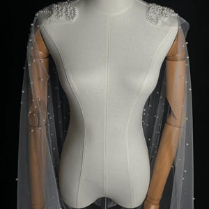 May include: A sheer white tulle capelet featuring scattered white pearls throughout the fabric. The shoulders are adorned with dense, decorative clusters of white pearls arranged in a floral-like pattern, extending into long, flowing panels.